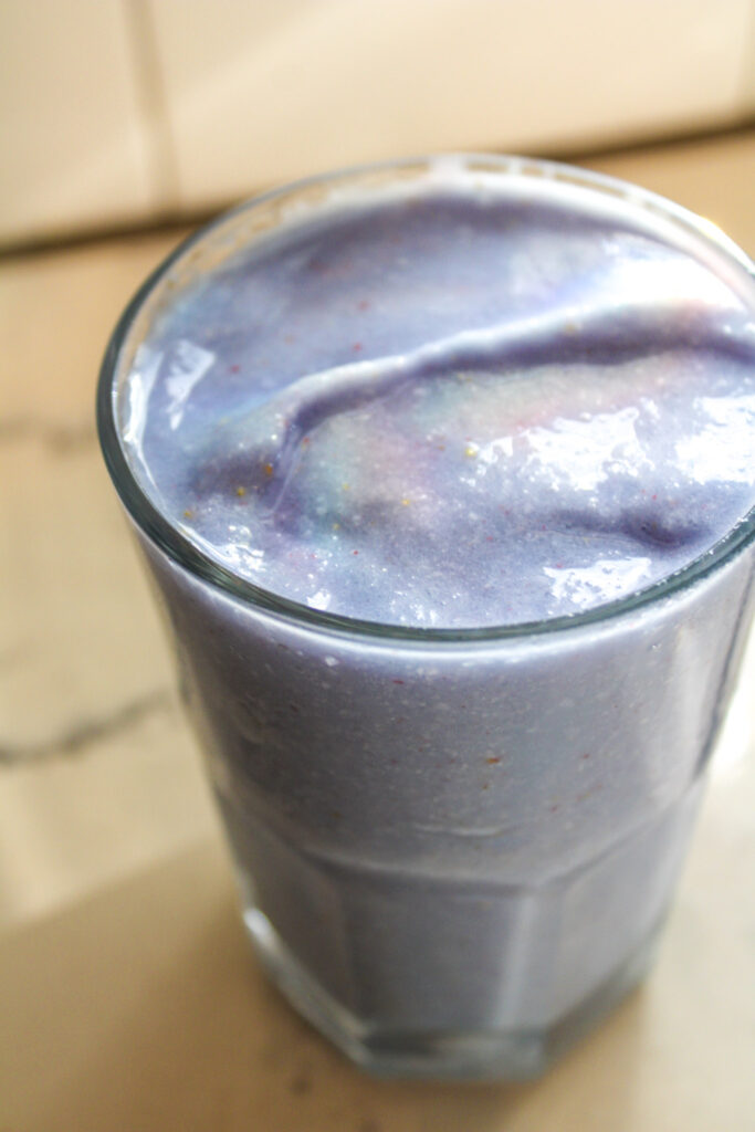 A lavender colored smoothie with blue spirulina, strawberries and coconut in a clear glass with a rainbow reflection over the top.