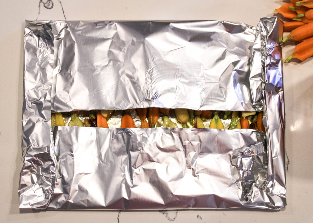 Carrots lined up in two rows on foil that's folded up over them with a little middle sliver opened in th emiddle of the foil where carrot tops are peakign out.