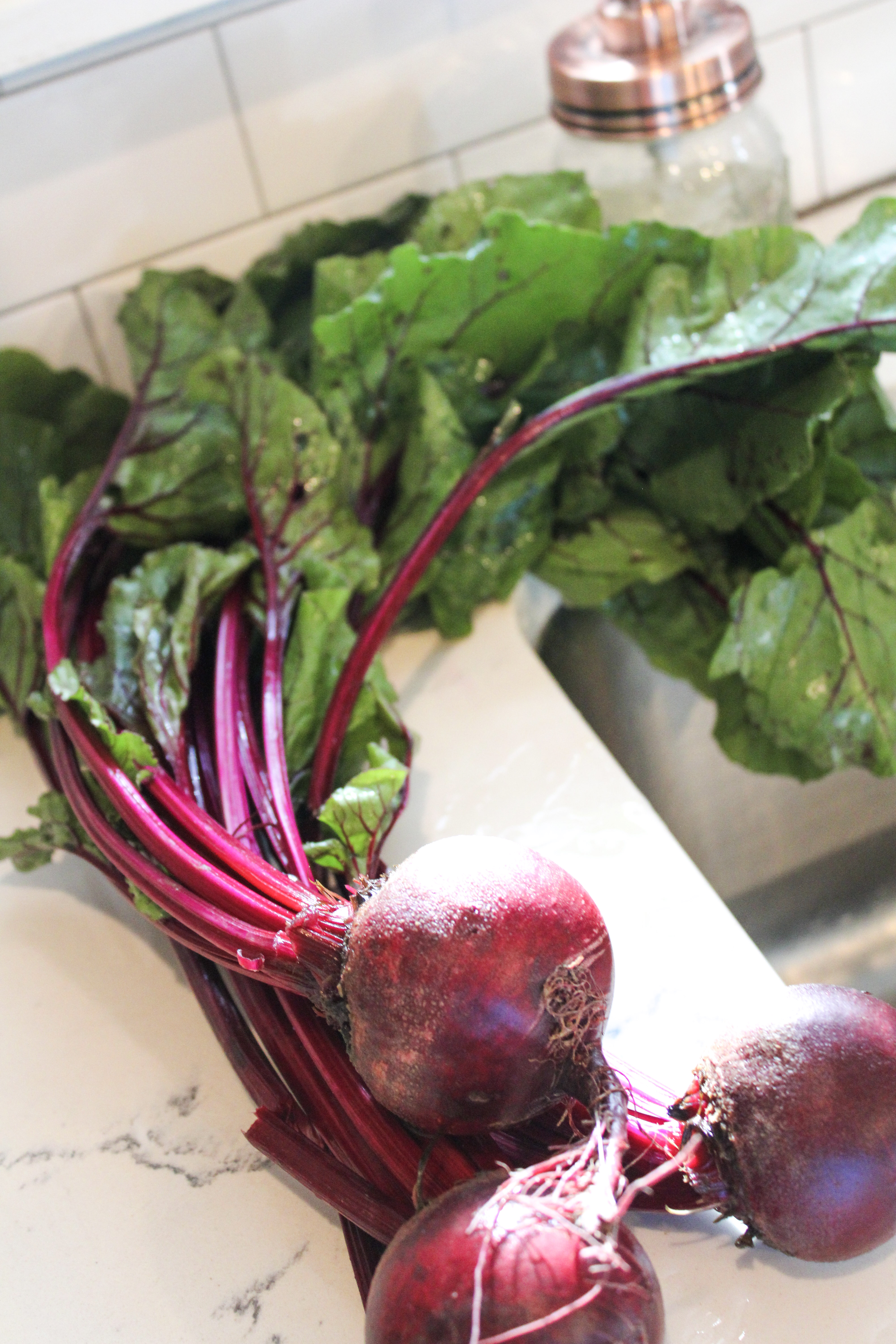 Entire ret beets by the sink on a white counter with their leaves and stems still attached.