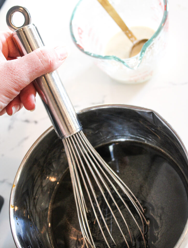 A hand whisking sauce with a cornstarch slurry in the background.