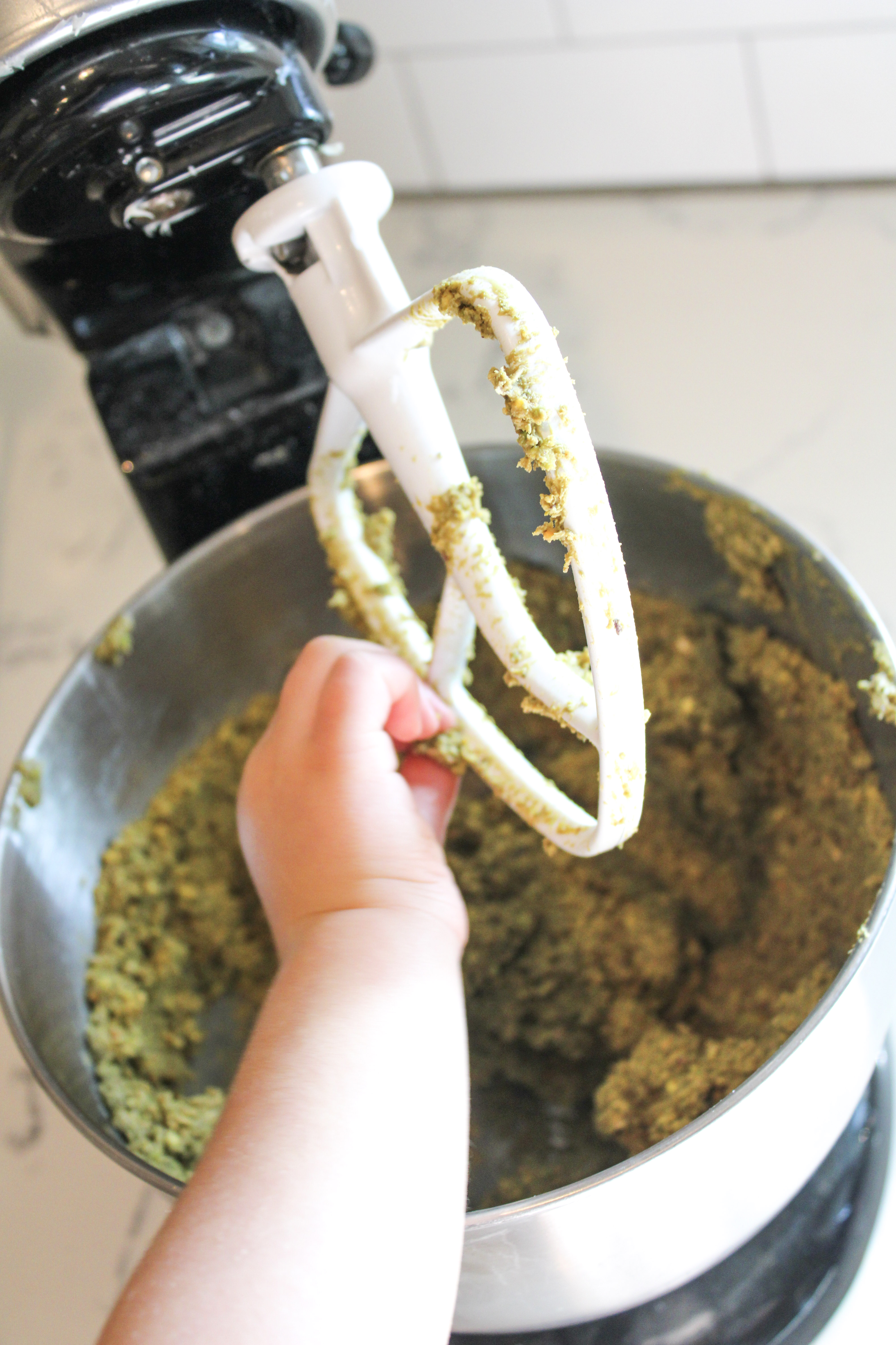 A stand mixer with the paddle sticking out that has pistachio bite dough on it and a toddler sneaking the dough with their hand.