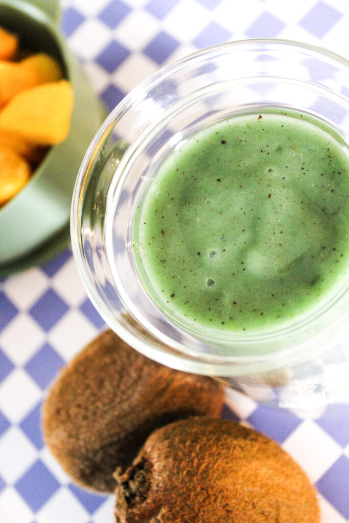 A green smoothie next to kiwis and mango on a purple checkered background.